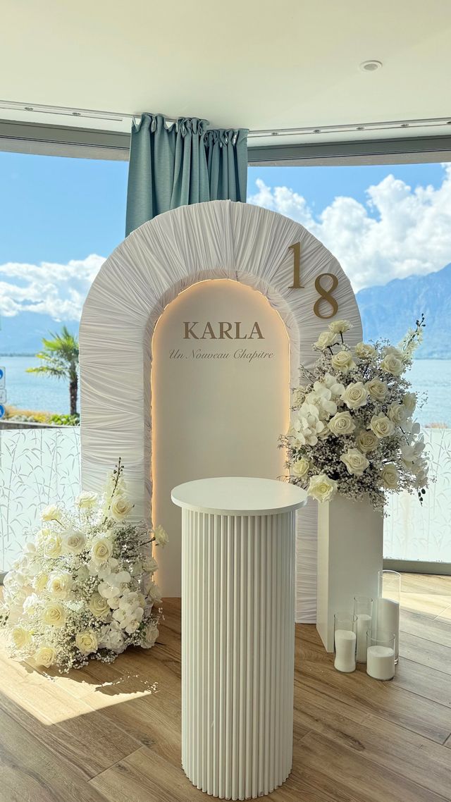 Décor d'anniversaire des 18 ans : arche drapée blanche avec lumière chaude, compositions de roses blanches et vue sur le lac